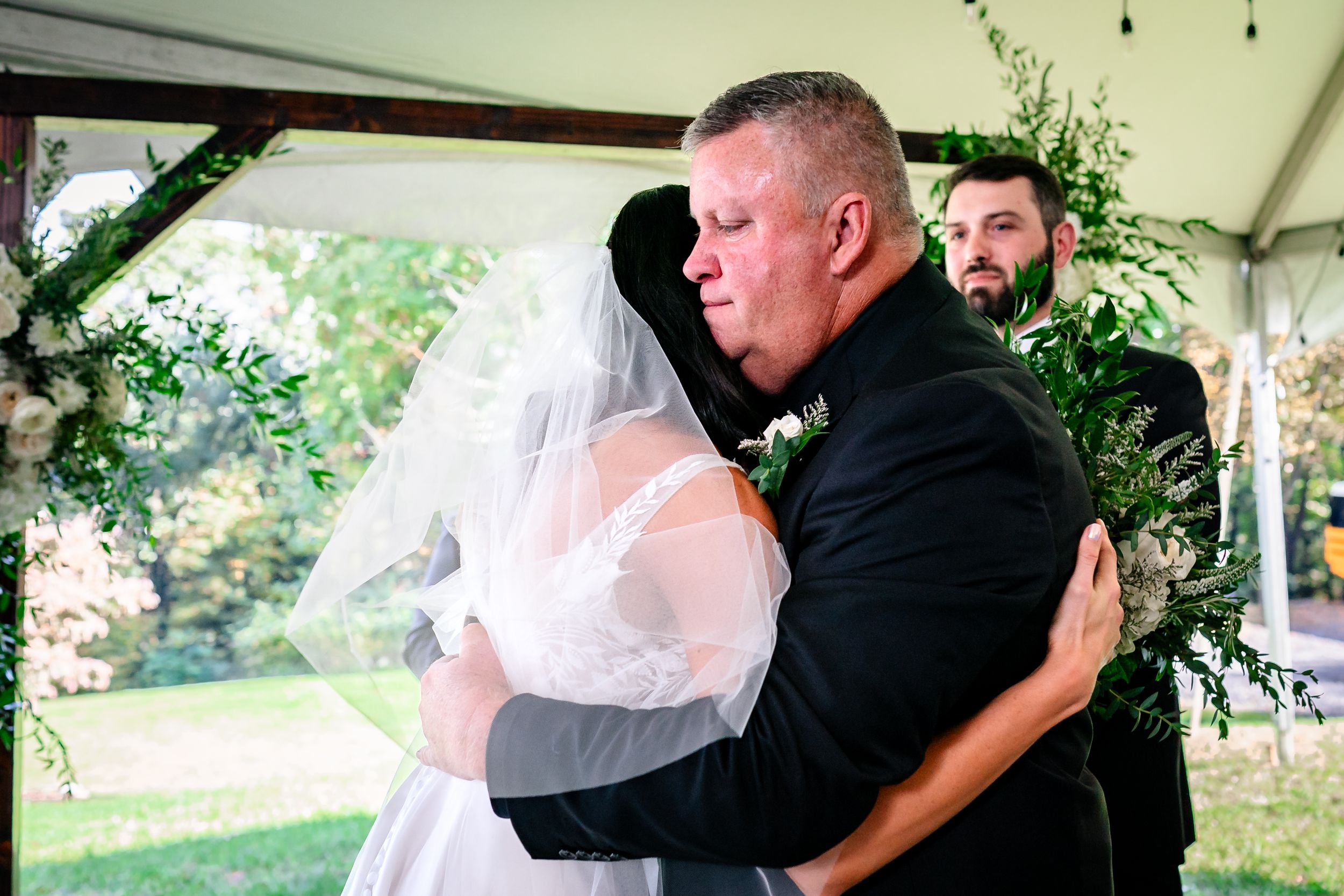 Emotional moment captured during outdoor wedding ceremony under tent with floral arch.