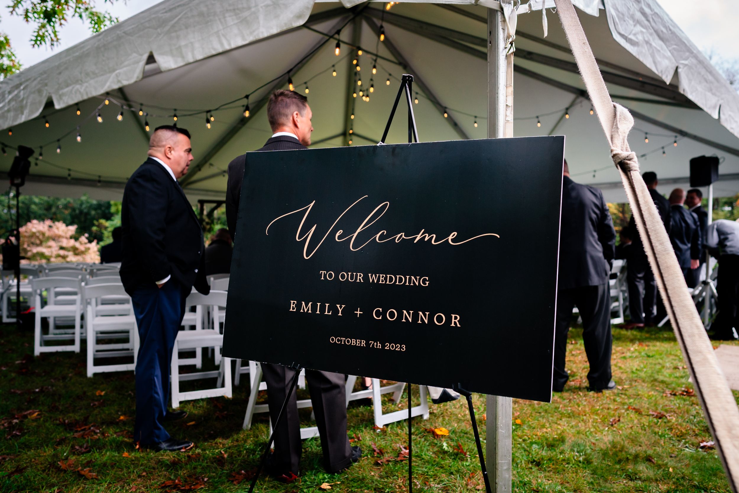 Welcome sign for wedding ceremony displayed under white tent with guests arriving.