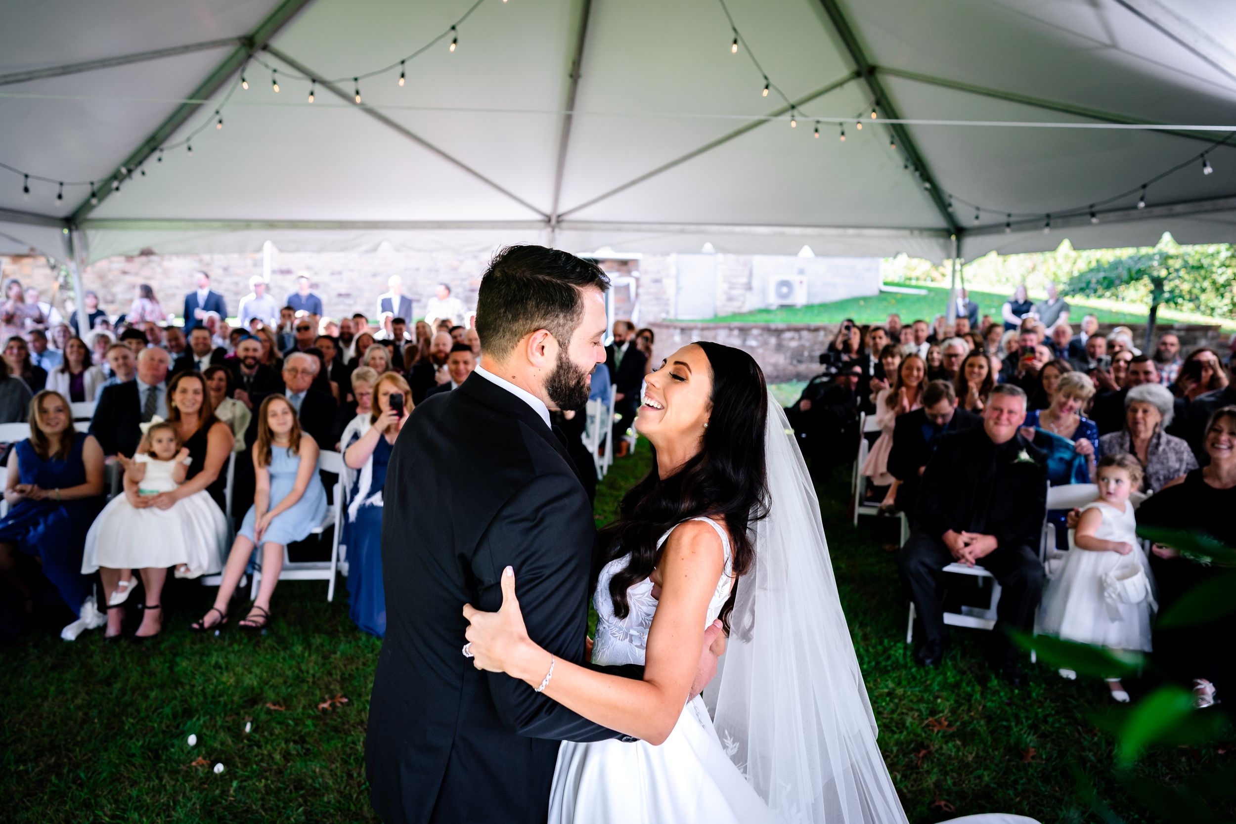 Wedding celebration under a white tent with string lights and gathered guests.