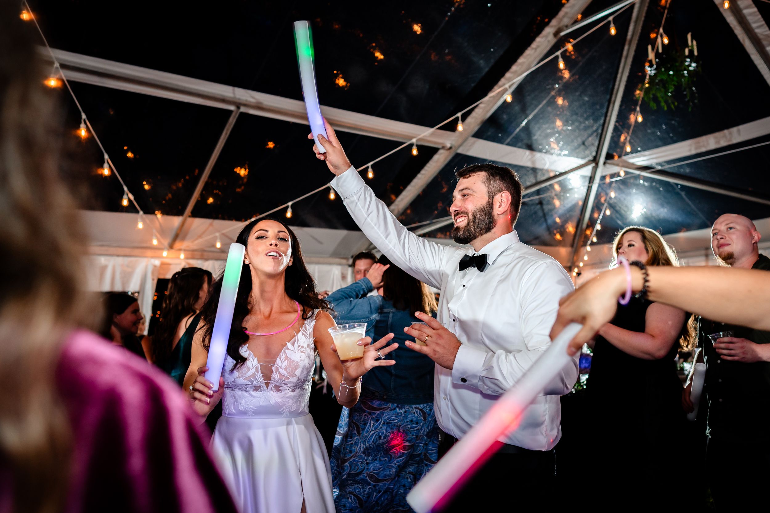 Guests dancing with colorful glow sticks under a clear tent at night.