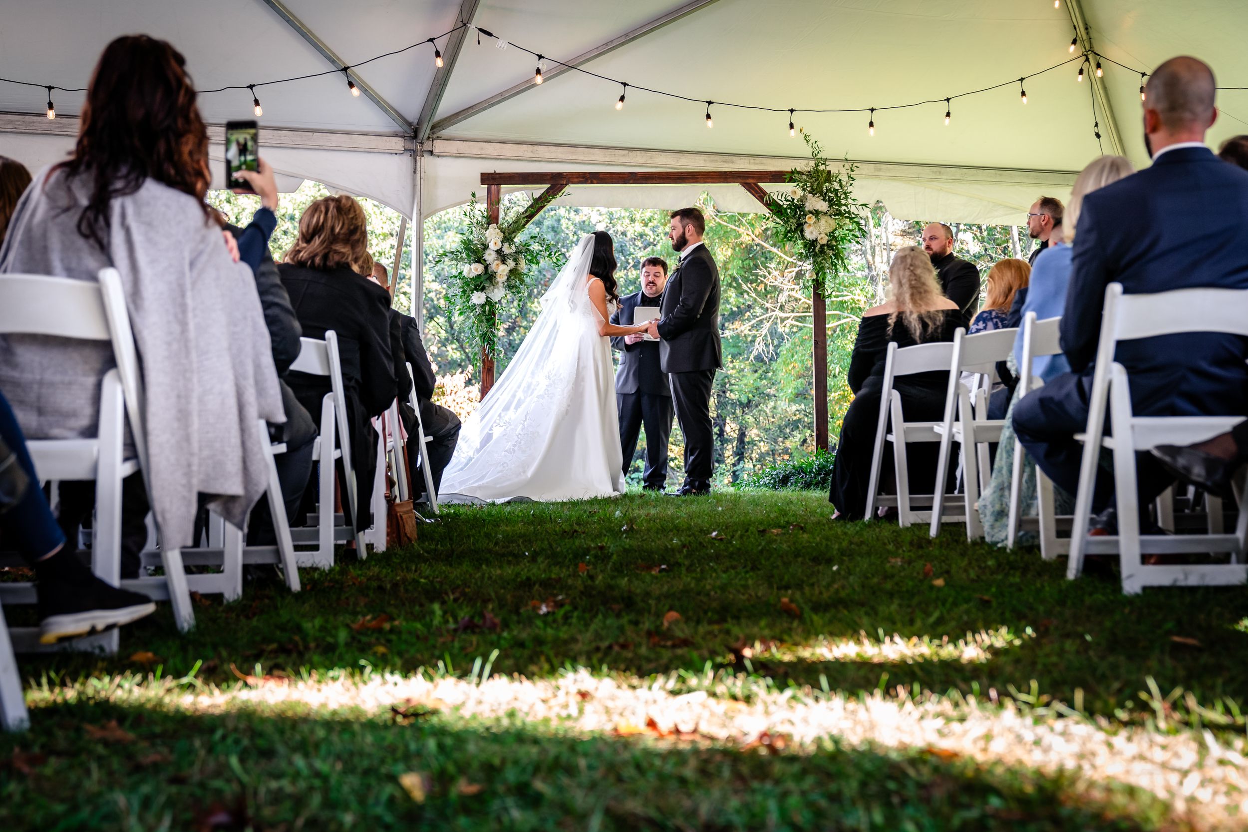 Outdoor tent wedding ceremony with guests seated on white chairs and sunlight streaming through.