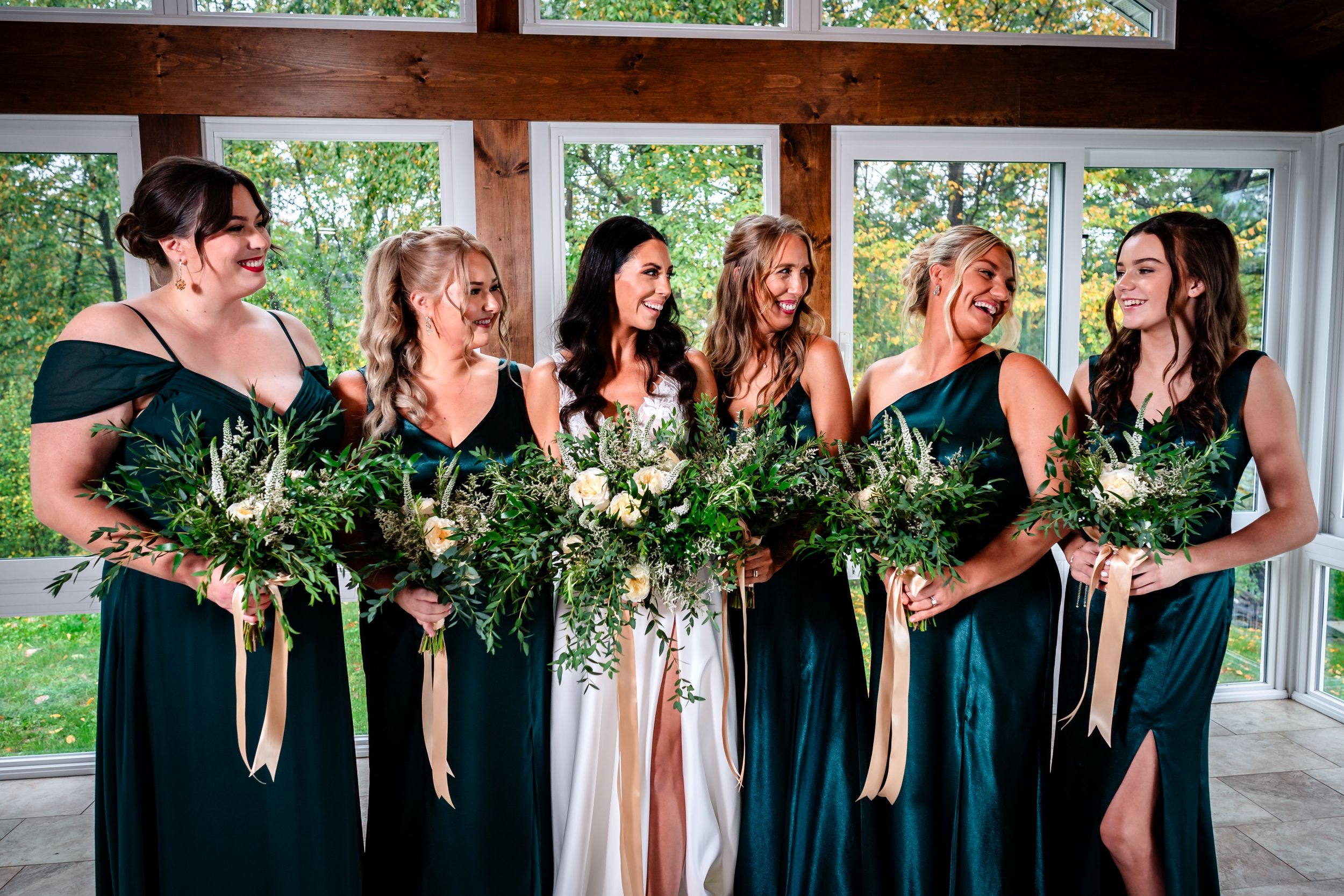Bridesmaids in elegant emerald gowns hold lush greenery bouquets while sharing a candid moment.