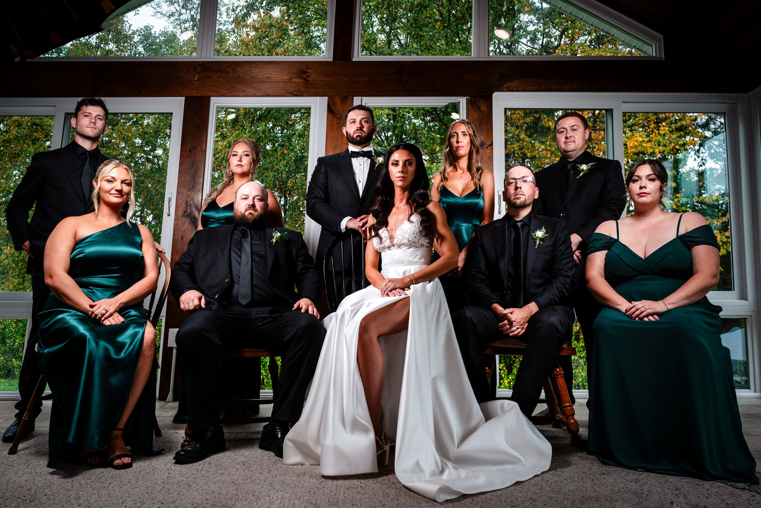 Wedding party poses together in formal attire with emerald green dresses and black suits in front of windows.