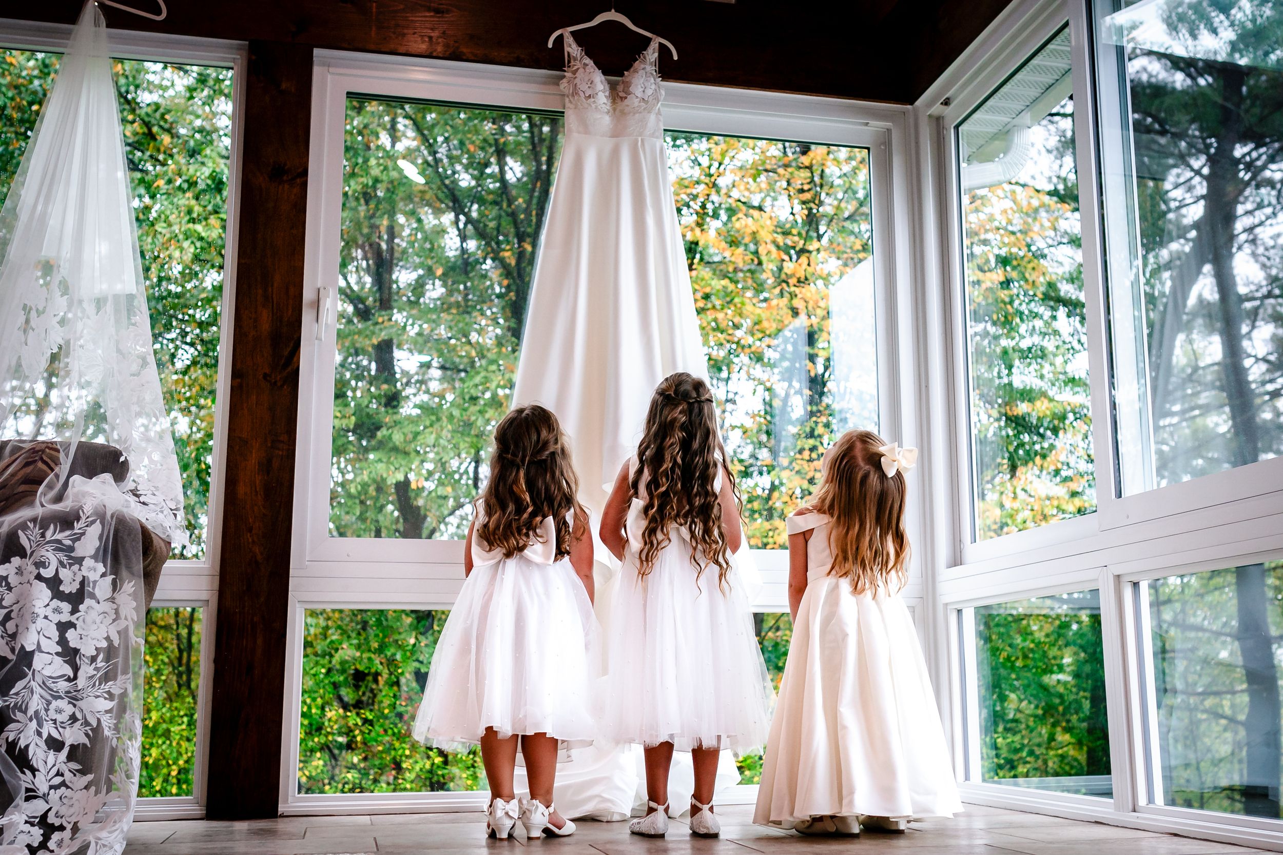 Multiple bridesmaids in white robes gathered by large windows during wedding preparations.