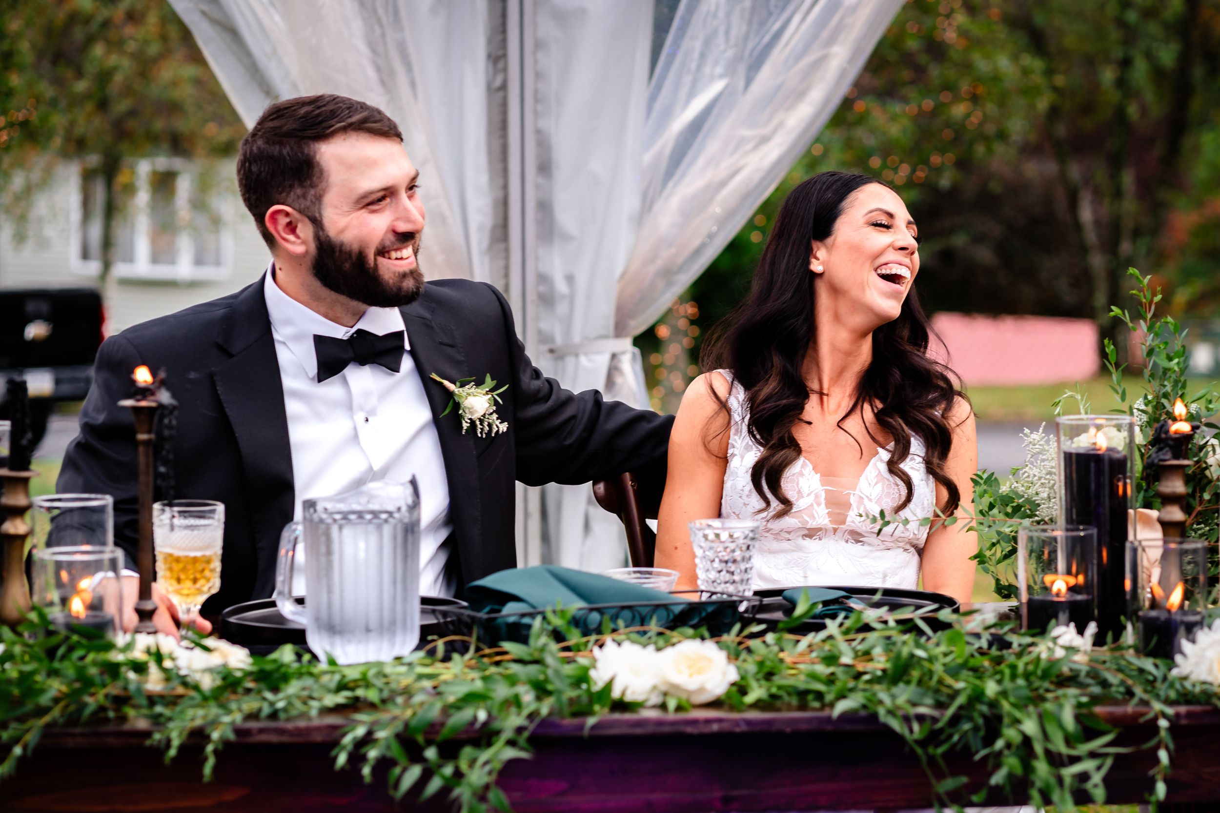 Wedding guests share laughter and conversation at an elegant outdoor reception under white drapes and greenery.