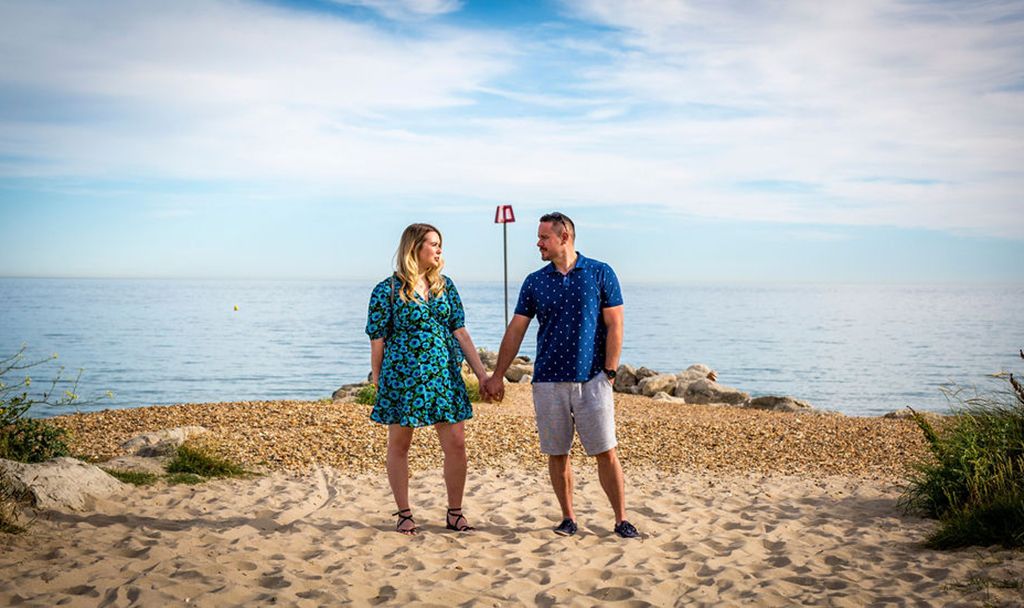 A couple poses on a sandy beach overlook with the ocean and blue sky in the background.