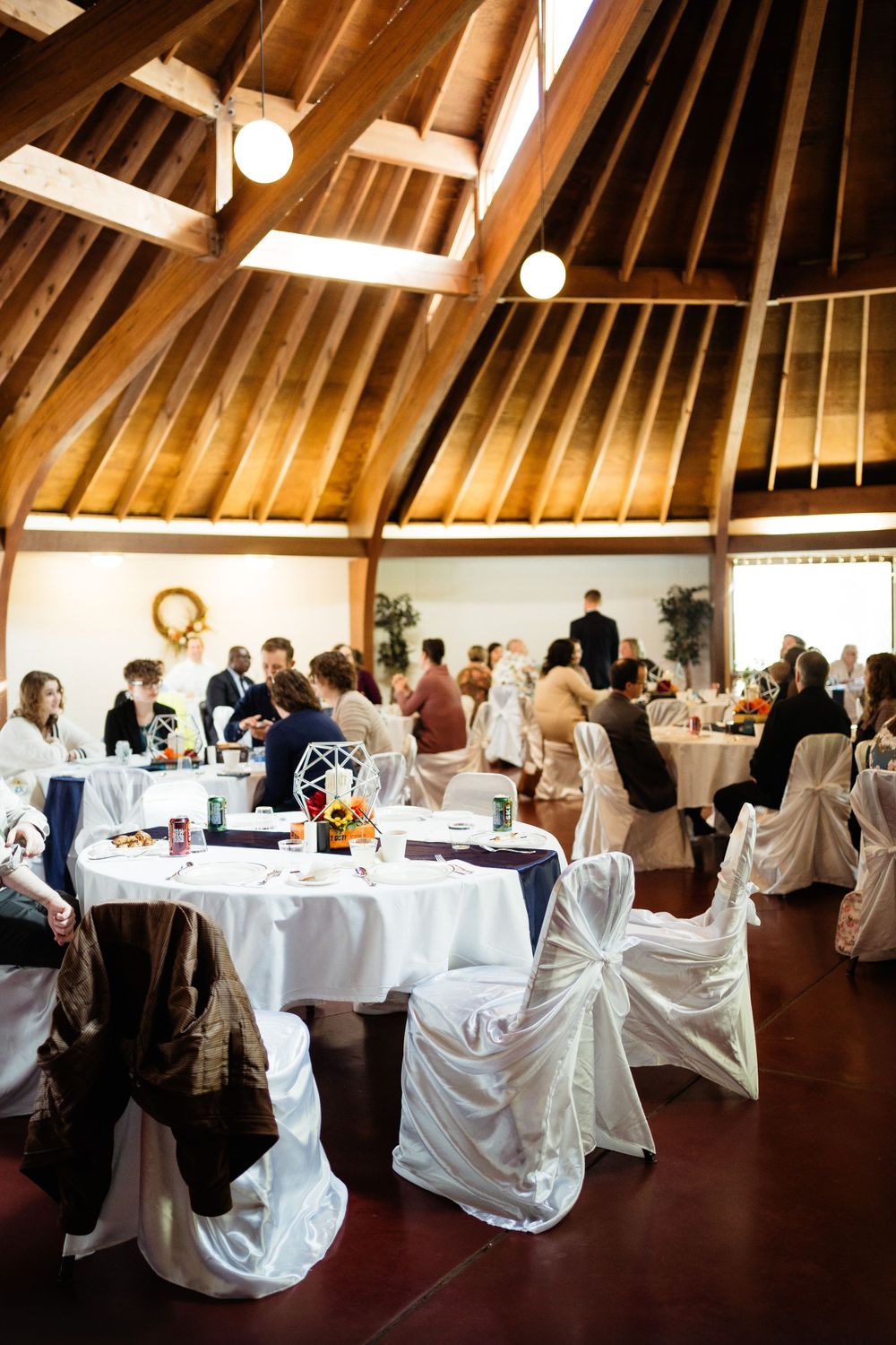 An elegant wedding reception setup in a wooden vaulted ceiling venue with white chair covers and table linens.