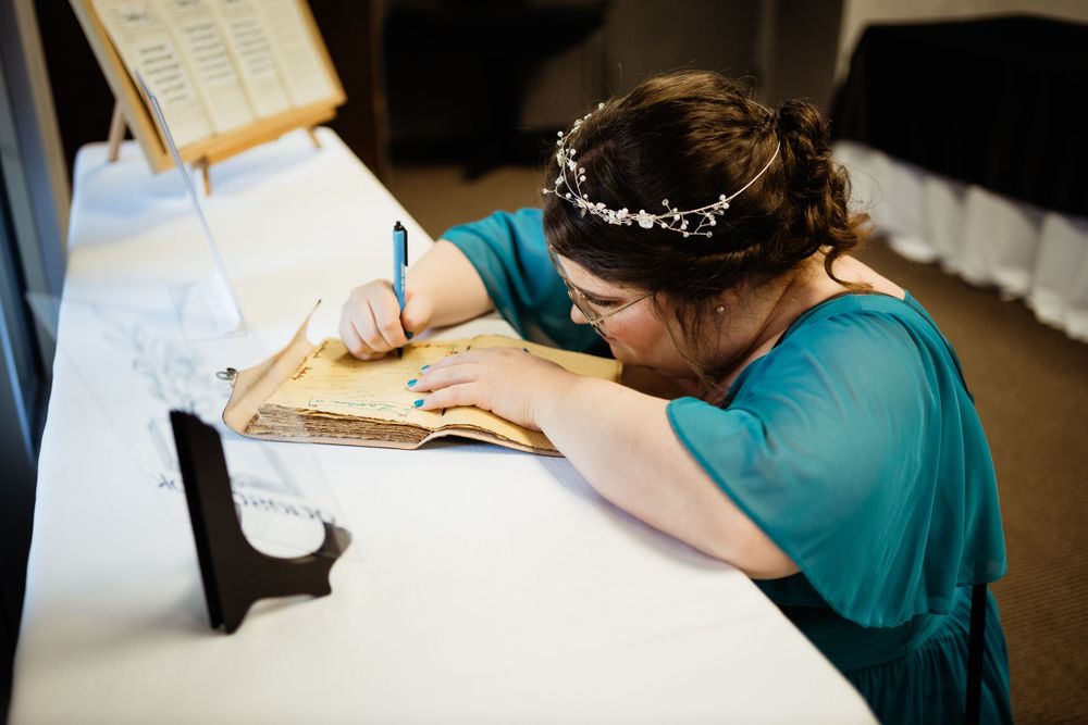 Someone in a teal dress signs a document at a desk during a wedding celebration.