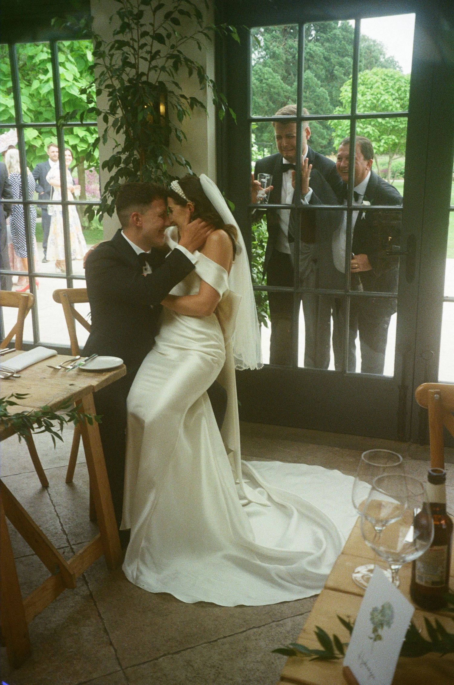 Newlyweds share romantic moment while guests watch through window.