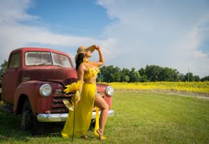 Yellow dress contrasts with rustic red truck in sunflower field during golden hour.