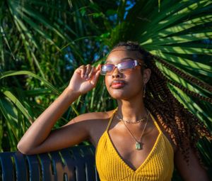 Person in a yellow top posed against tropical palm fronds in bright sunlight.