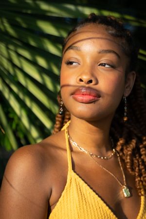 Close up portrait of someone wearing a yellow top and jewelry against palm leaves.