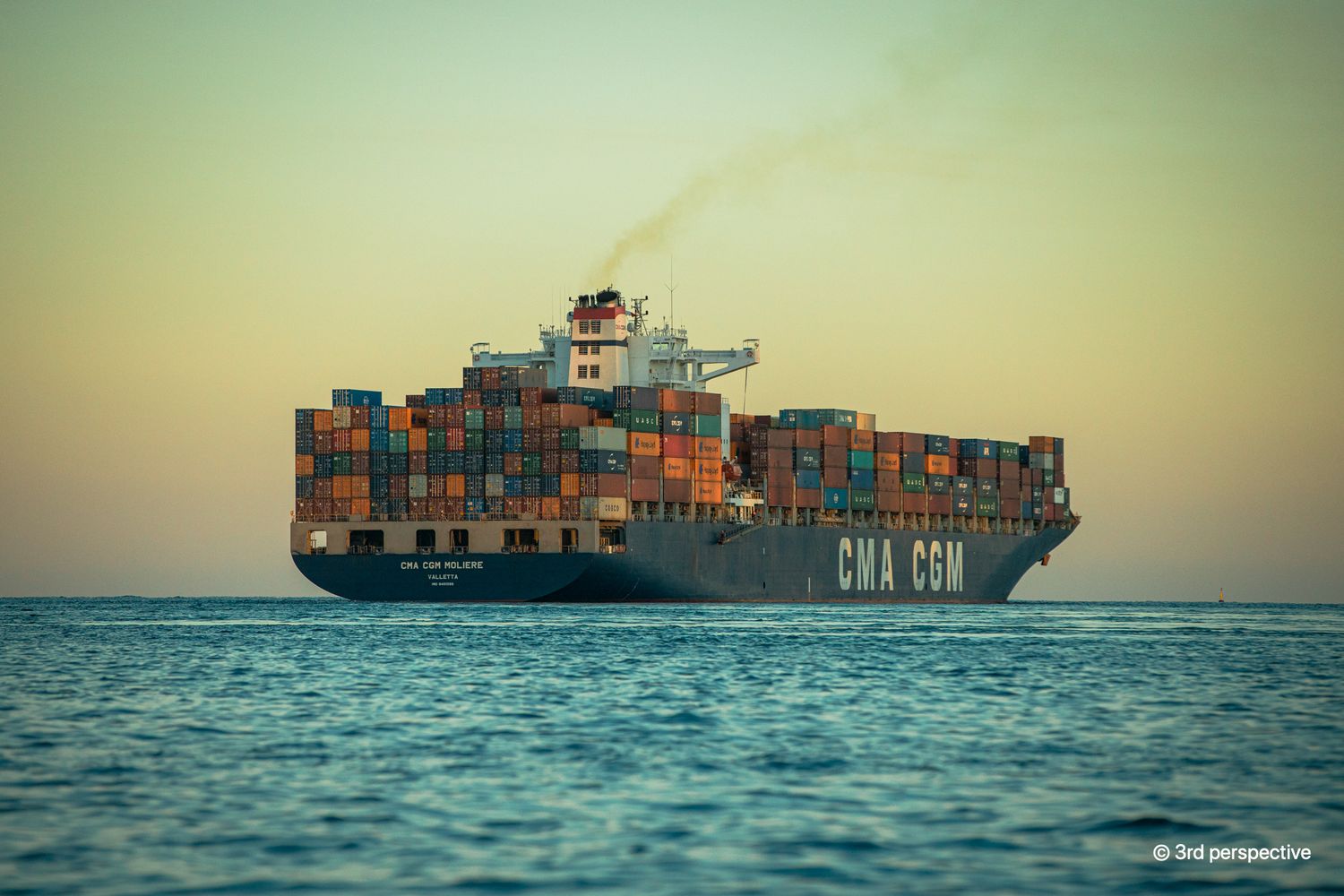 Large CMA CGM container cargo ship loaded with colorful shipping containers sailing on calm ocean at sunset.