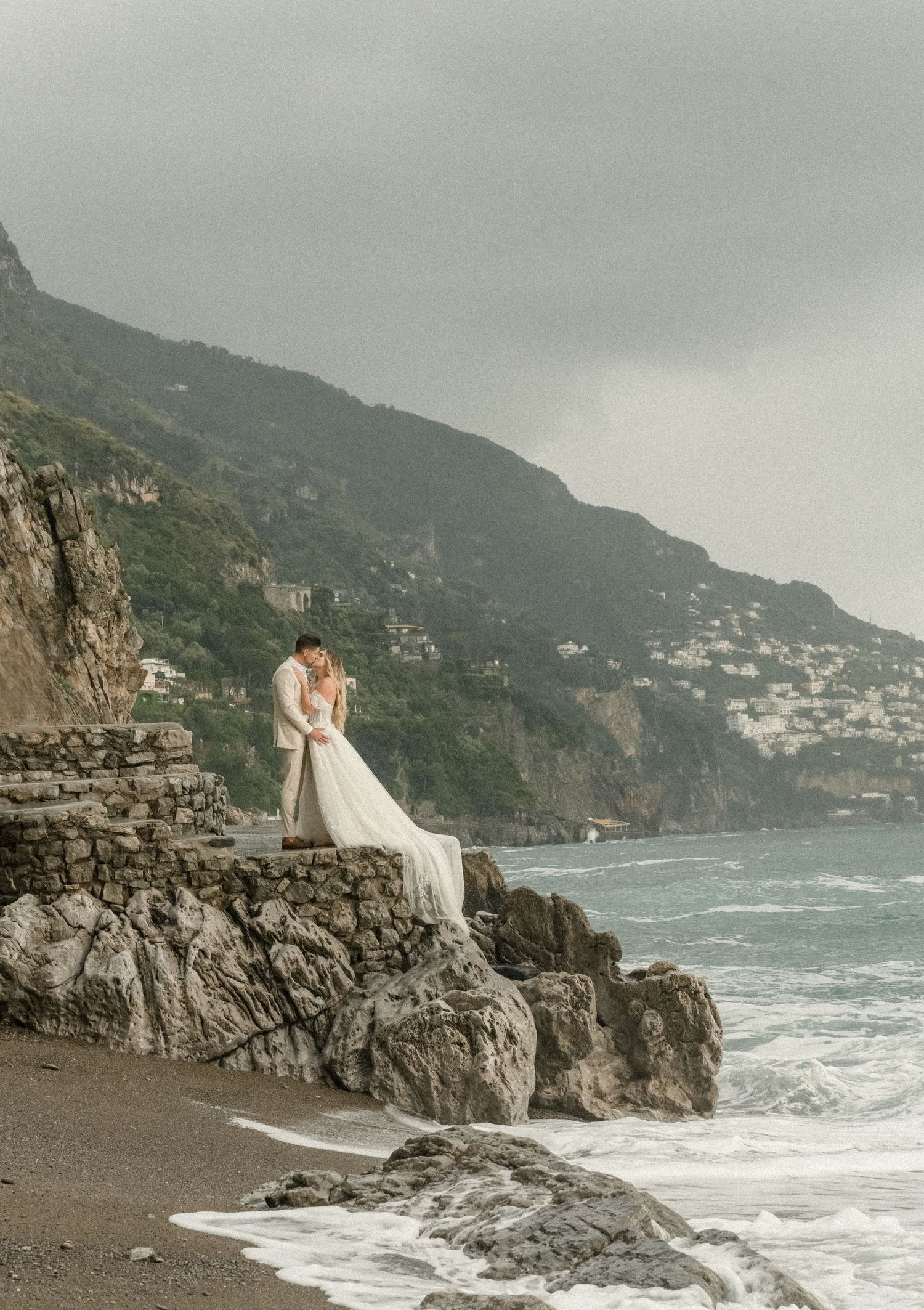 Wedding couple sitting on coastal rocks overlooking Mediterranean Sea.