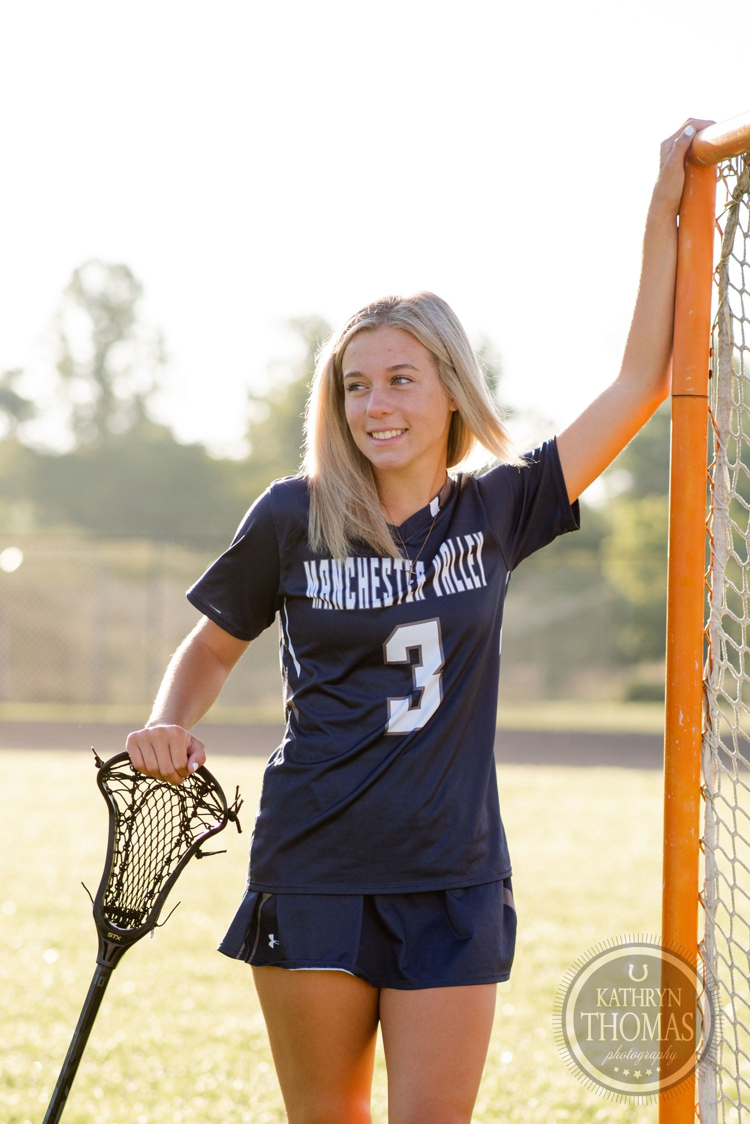 Lacrosse player in uniform poses with stick next to goal post during golden hour.