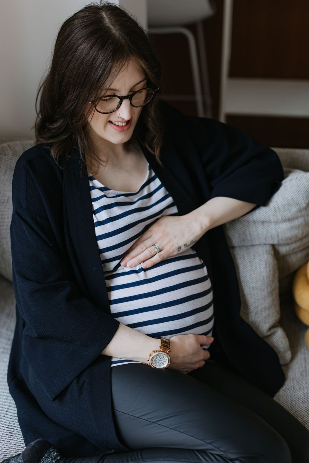 Pregnant woman in striped shirt and black cardigan sitting on couch holding her belly while smiling.