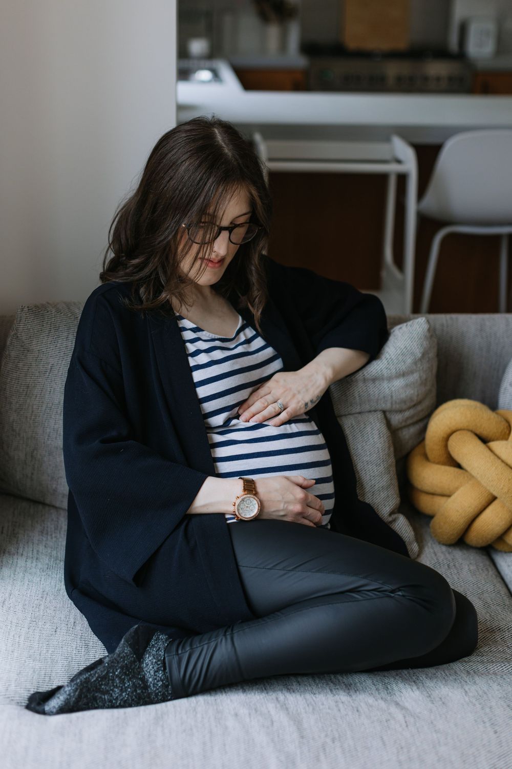 Someone wearing a navy blazer and striped shirt sits on a gray couch with a yellow knot pillow, touching their midsection.