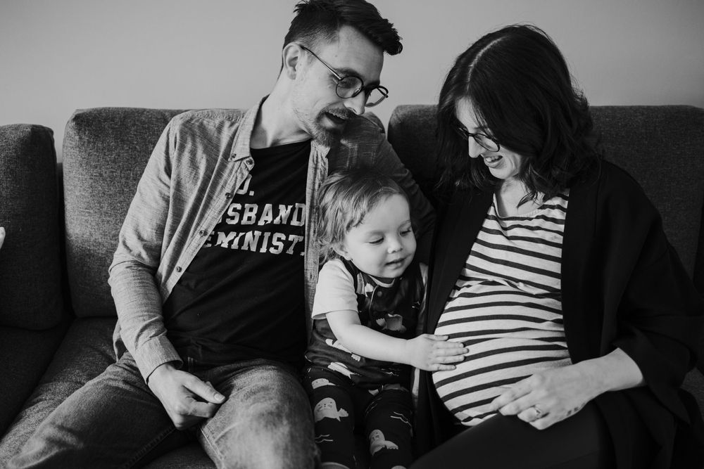Black and white intimate family photo of couple and young child sitting together on a couch reading a book.