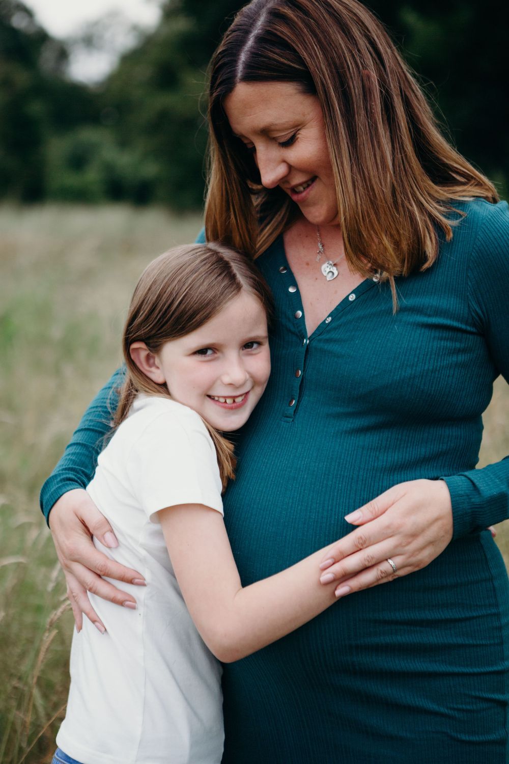 A tender moment between a pregnant woman in a teal dress and a young child embracing outdoors in a field.