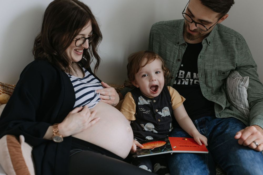 A family sits together reading while a toddler expresses excitement on a couch.
