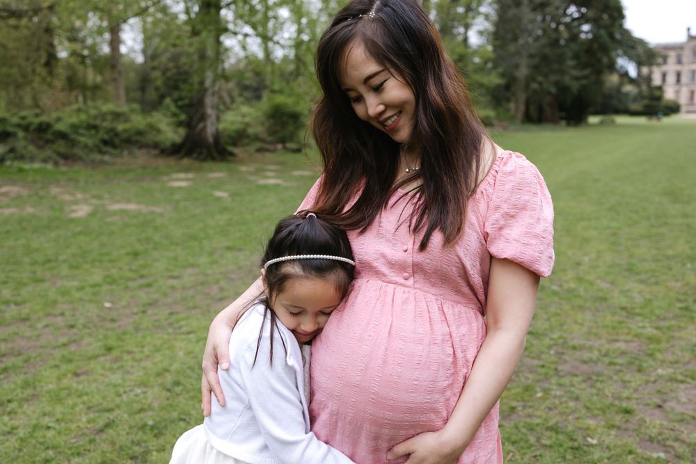 A mother in a pink maternity dress stands with her child in a grassy field while gently touching her pregnant belly.
