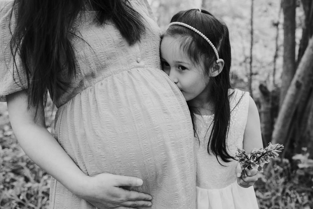 A tender black and white maternity moment as a small child lovingly leans close to a pregnant belly in a natural setting.