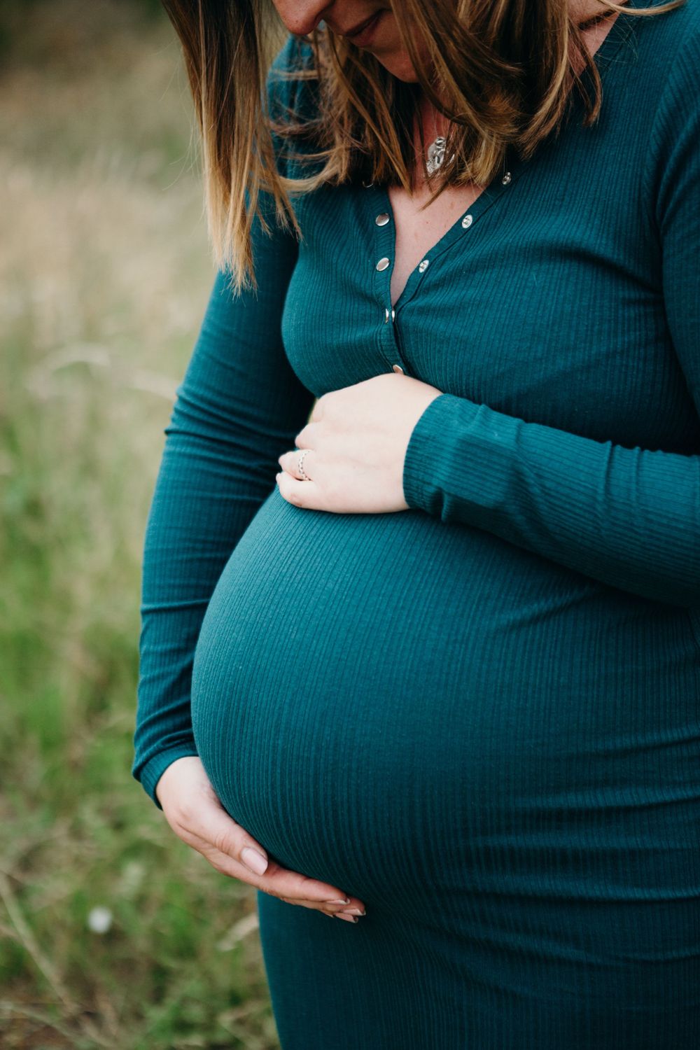 A pregnant figure in a teal ribbed dress cradles her baby bump while standing in a grassy outdoor setting.