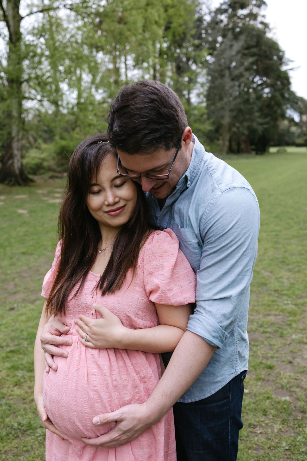 A couple stands outdoors in a tender embrace, with the woman wearing a pink dress showing her pregnancy belly.