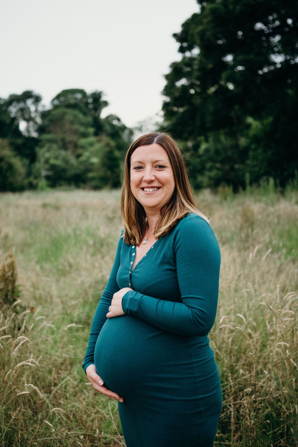 Expectant mother in teal dress stands in rustic field with natural foliage background during maternity photoshoot.