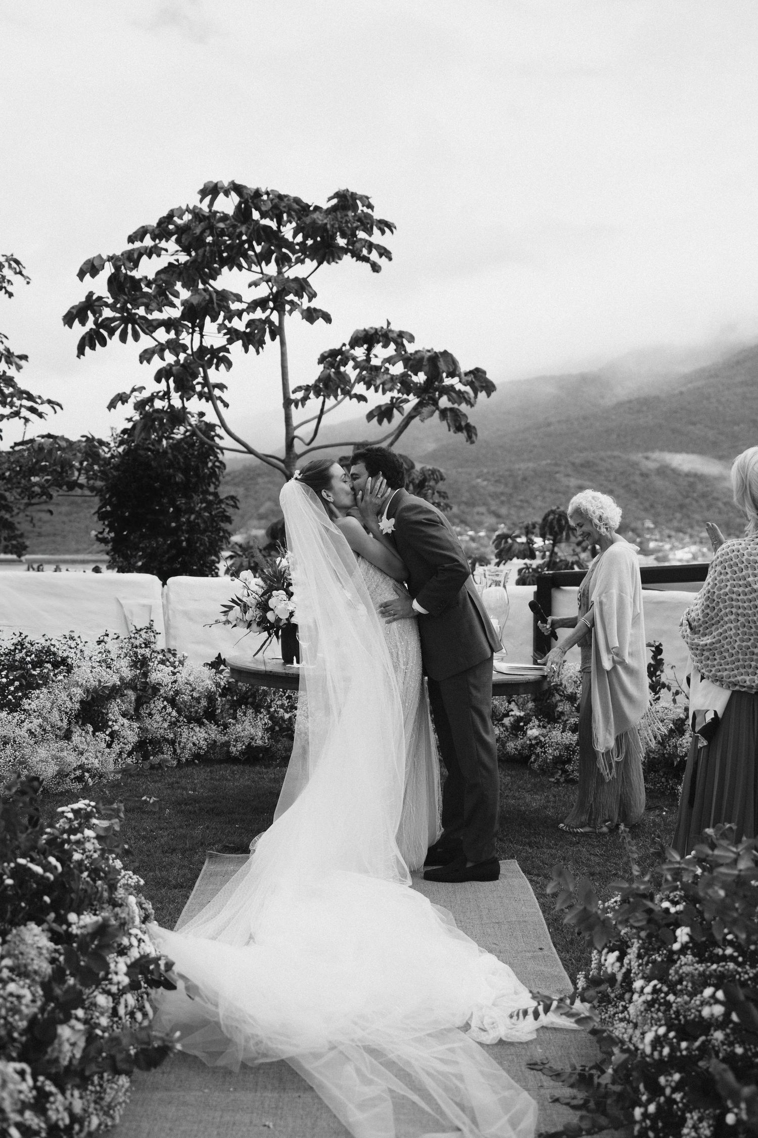 Black and white photograph of couple embracing during wedding ceremony in scenic mountain garden.