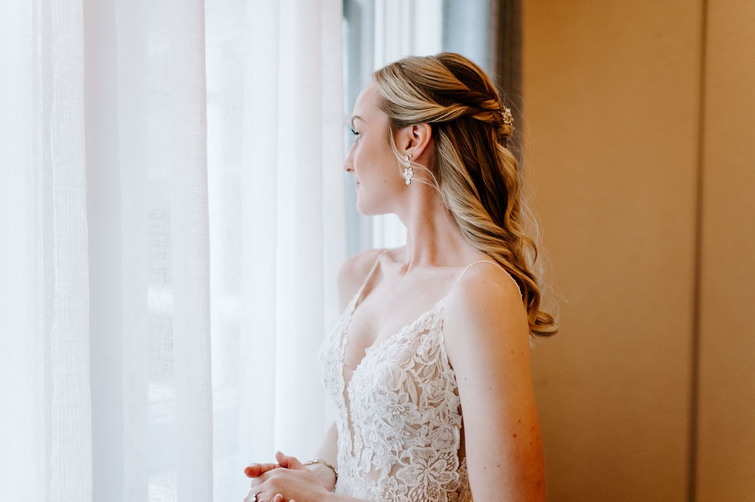 Bride in delicate lace wedding dress puts on earrings while getting ready by window light.