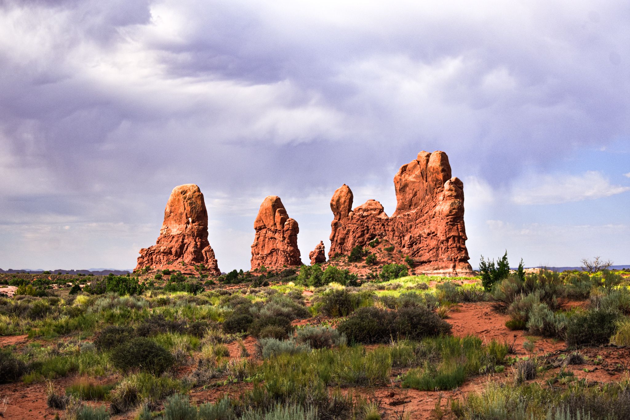 Majestic red sandstone rock formations rise from the desert floor against a cloudy purple sky.