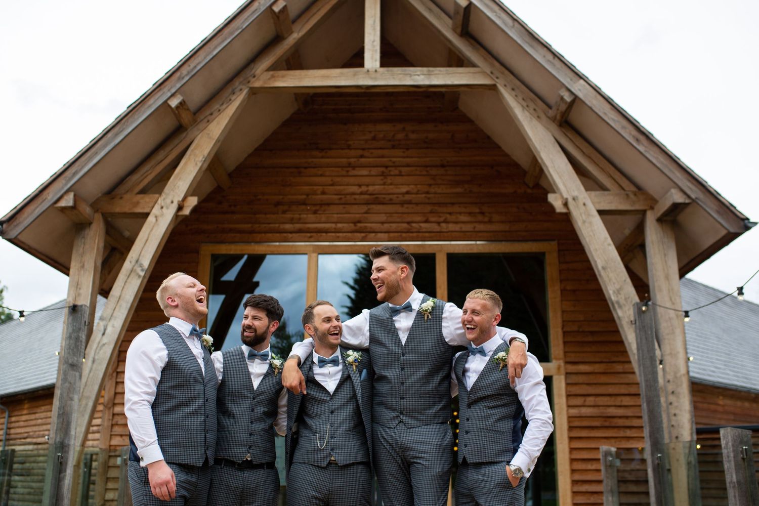 Groomsmen posing together in matching vests outside a rustic wooden venue.
