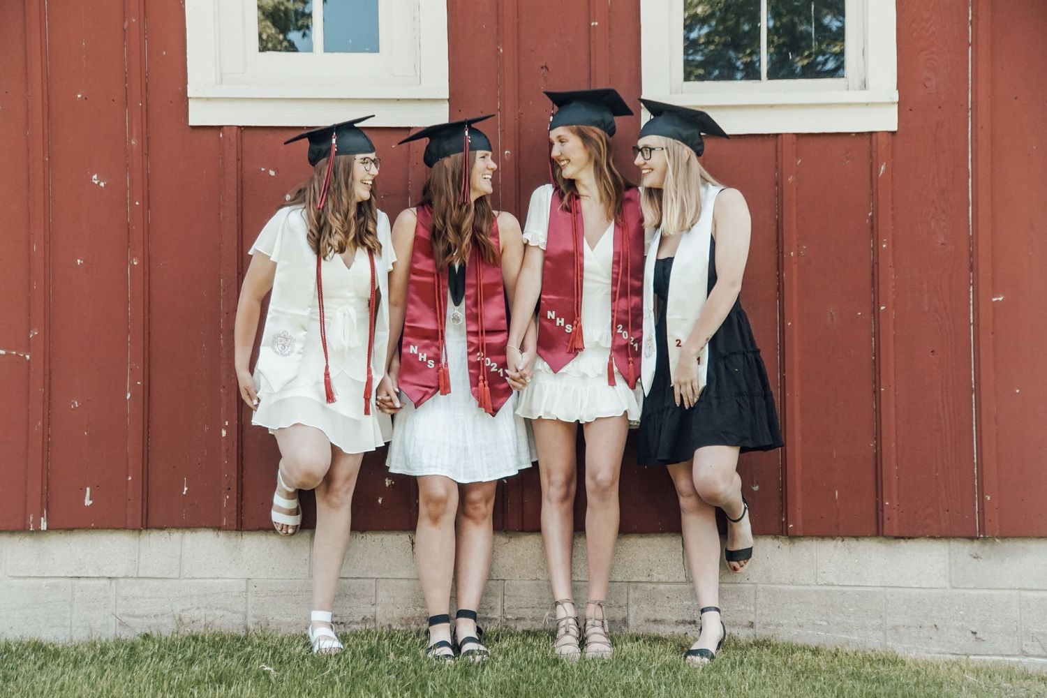 Group of graduates in white and black dresses with caps posing together against a red barn wall.