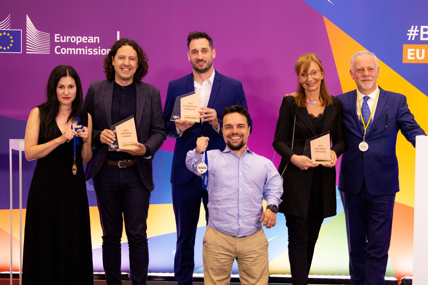 Award winners pose on stage at a European Commission ceremony with colorful backdrop.