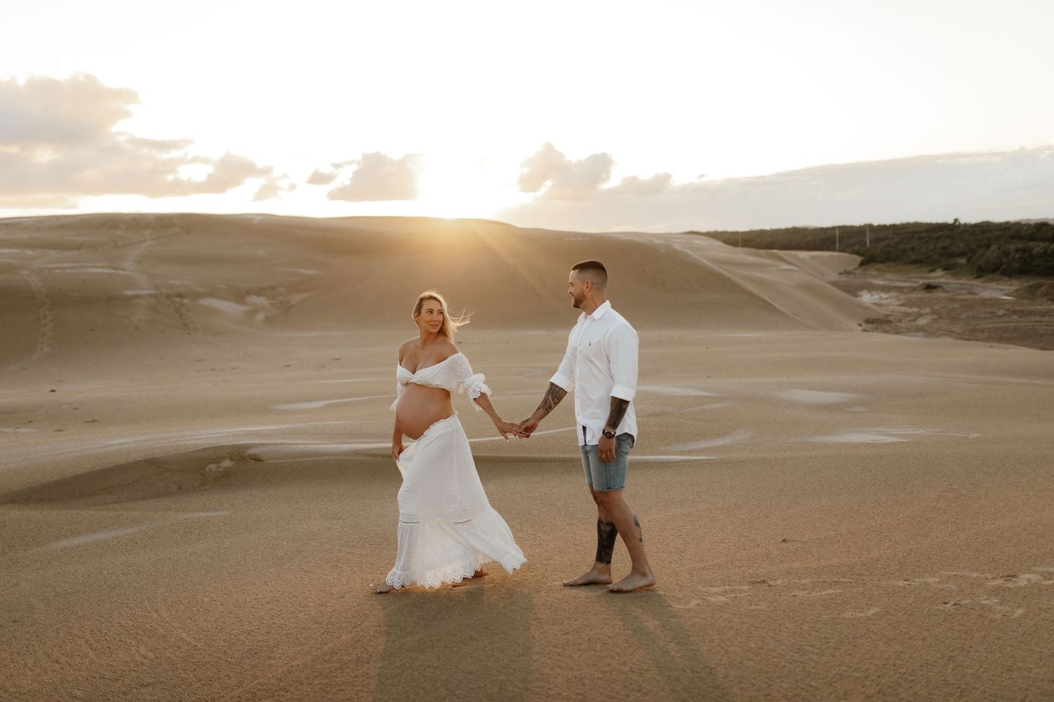 Romantic desert maternity photo session at sunset with couple holding hands on sand dunes.
