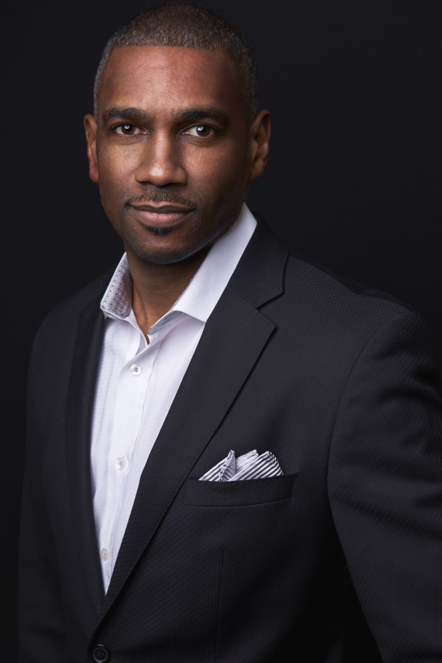 A well-dressed man in a black suit with pocket square poses confidently against a dark studio background.