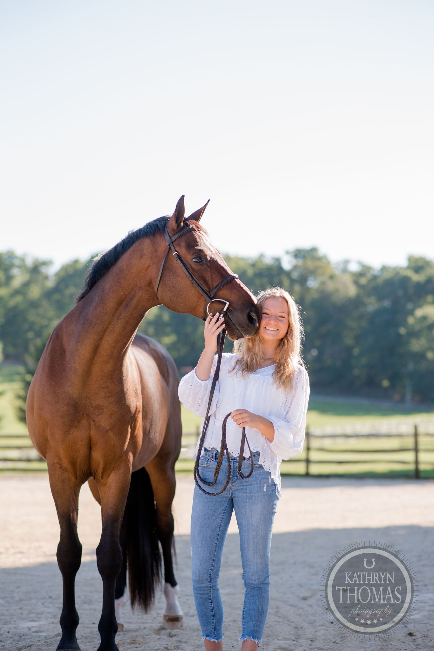 Bay horse and casual dressed person posing together in sandy arena during golden hour.