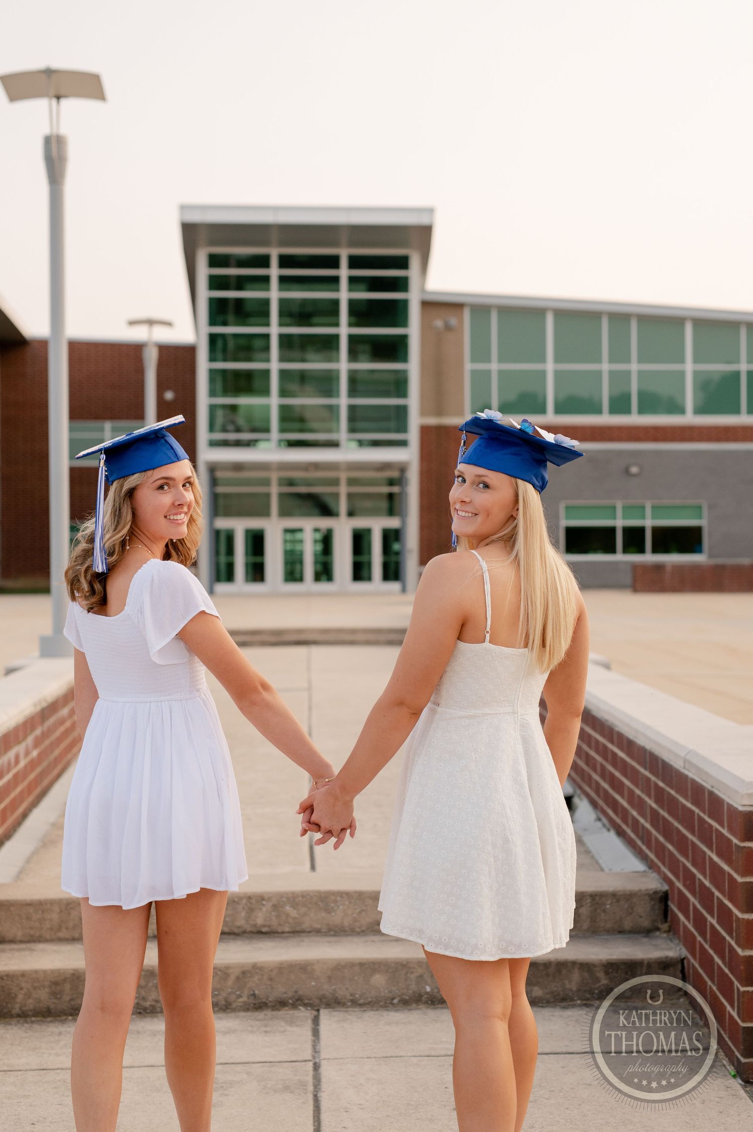 Two graduates in white dresses and blue caps holding hands in front of a modern school building.