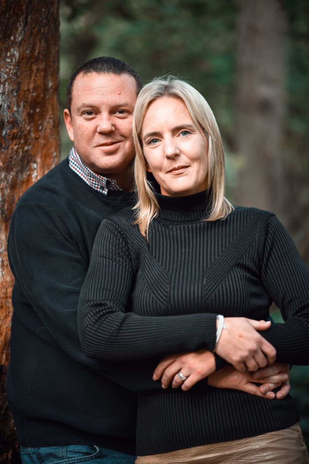 An intimate portrait of a couple in dark sweaters standing by a tree in the woods.