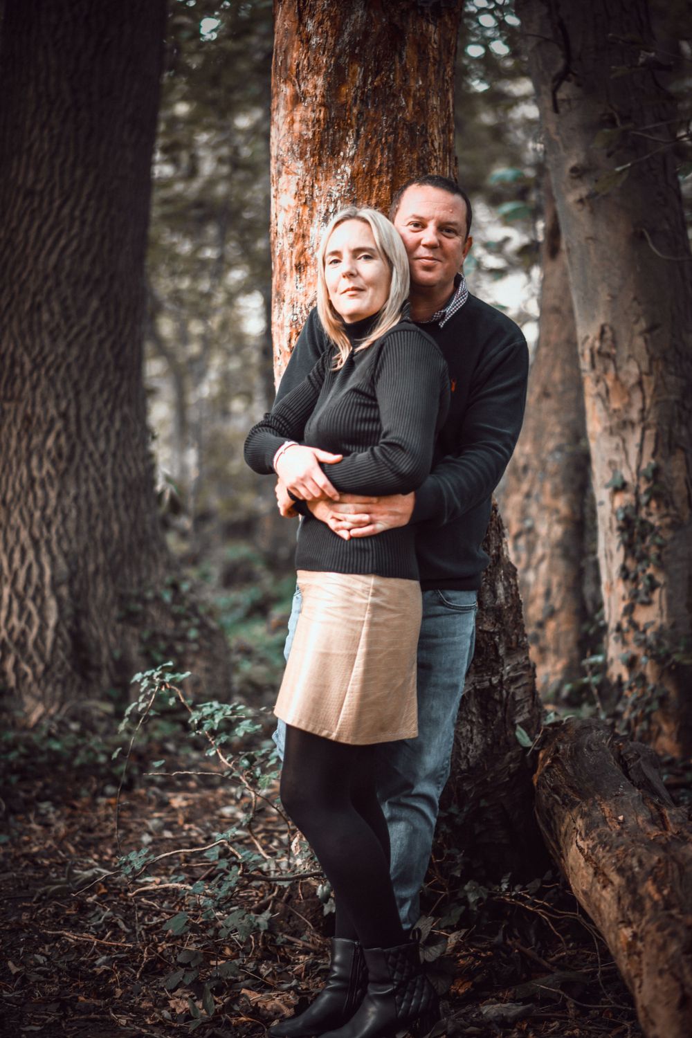 A loving couple poses together against a tree in a wooded setting wearing dark clothing.