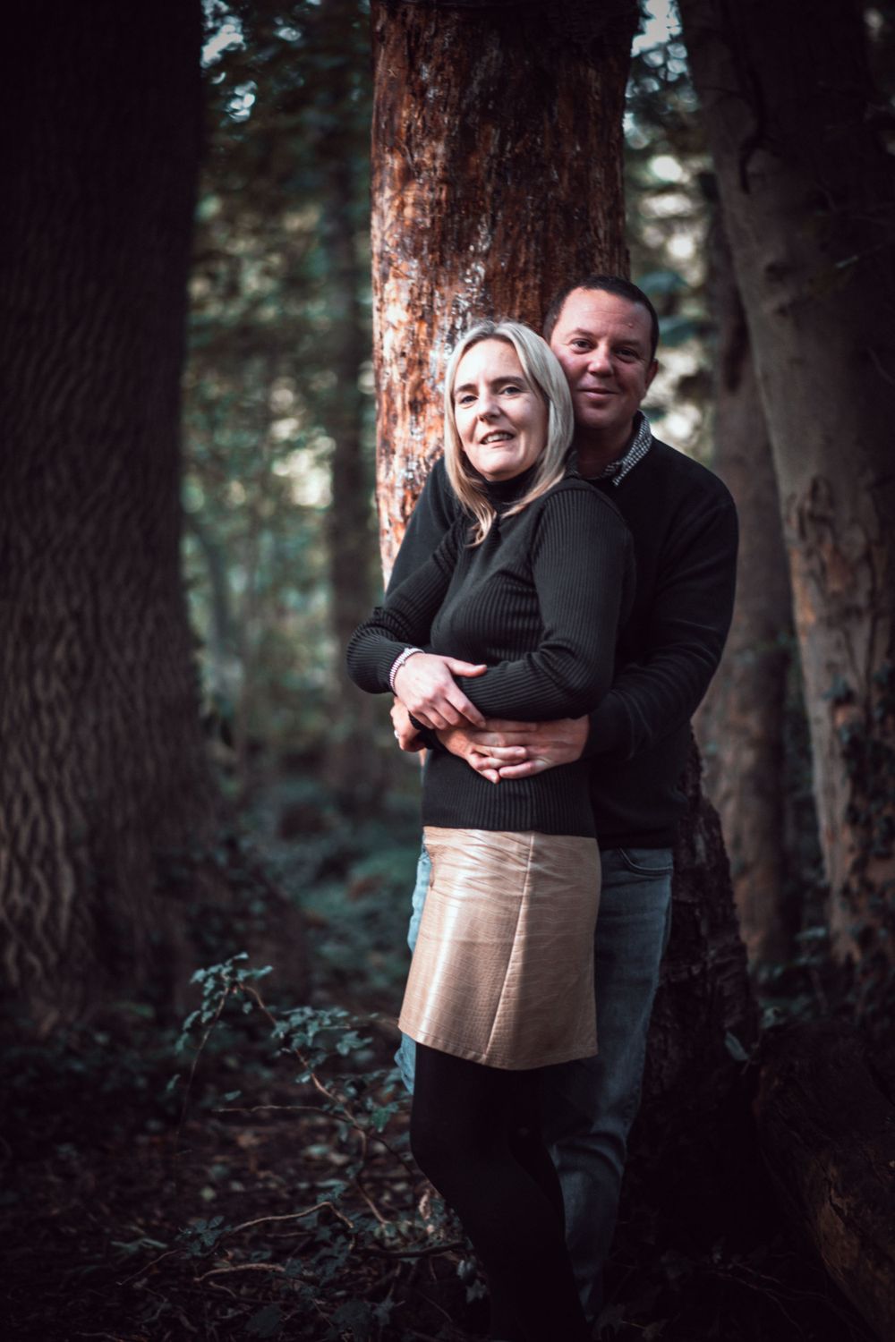 A romantic forest portrait of a couple embracing against a tree trunk in moody lighting.