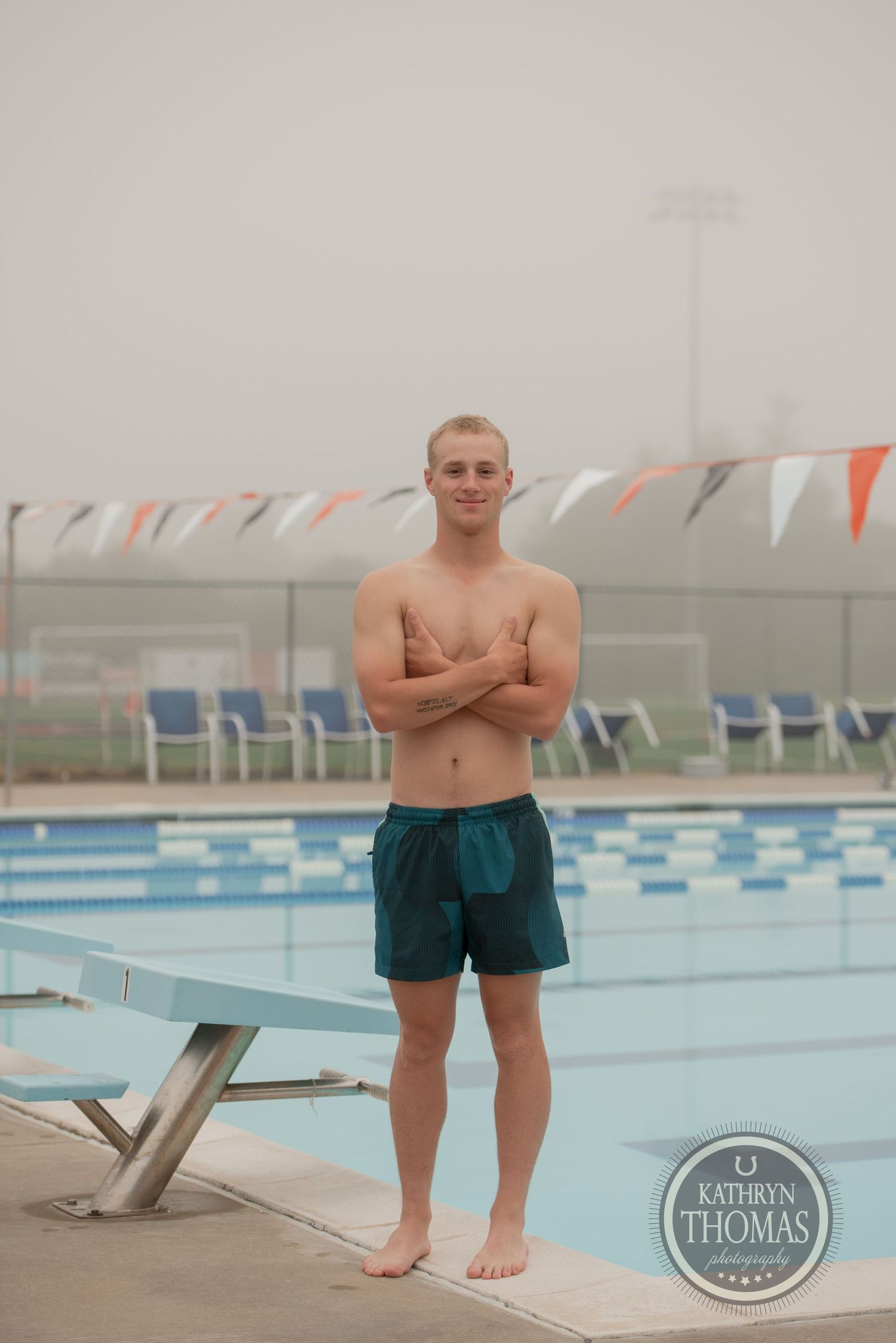 A swimmer stands on diving platform with arms crossed and pool with bunting flags visible.