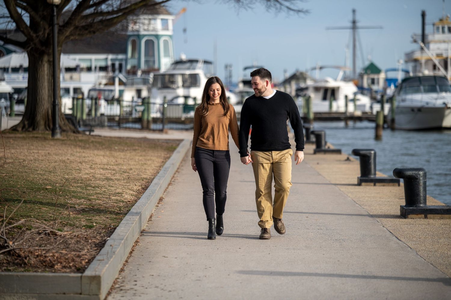 Couple walking together along waterfront boardwalk with boats and marina in background.