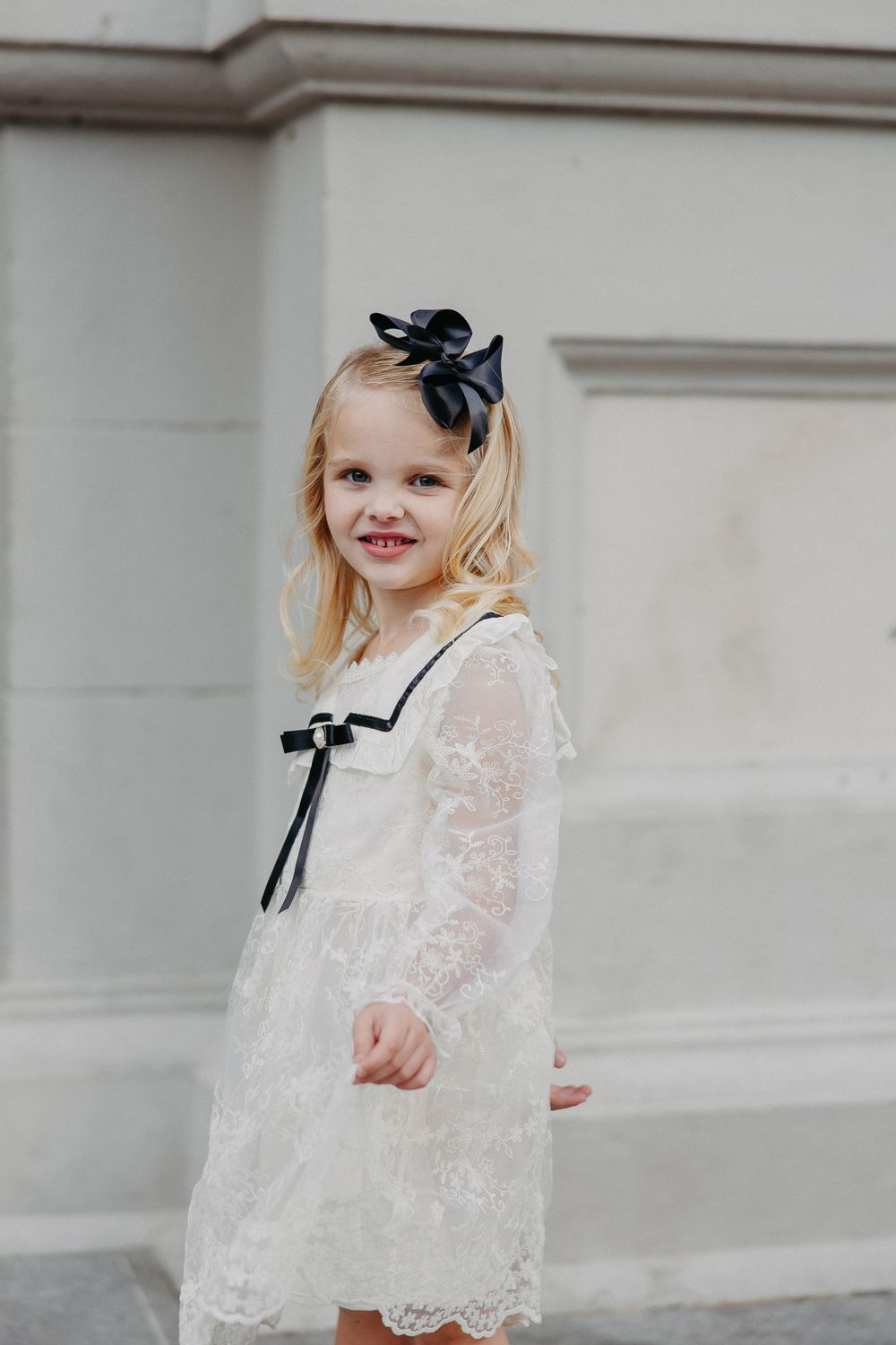 A young girl in a white lace dress with a black bow wearing a sweet smile.