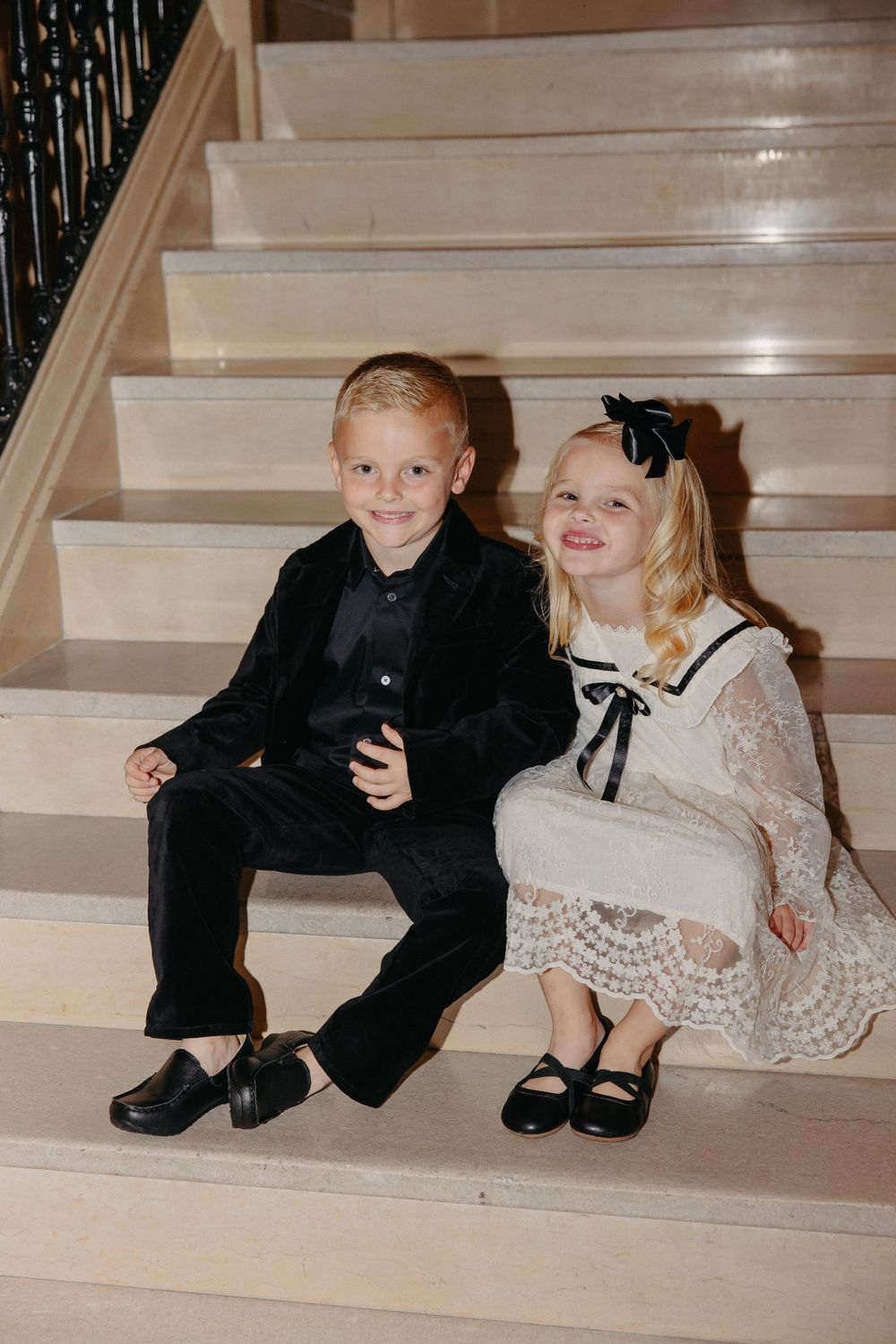 Two children in formal attire sitting together on elegant white stairs.