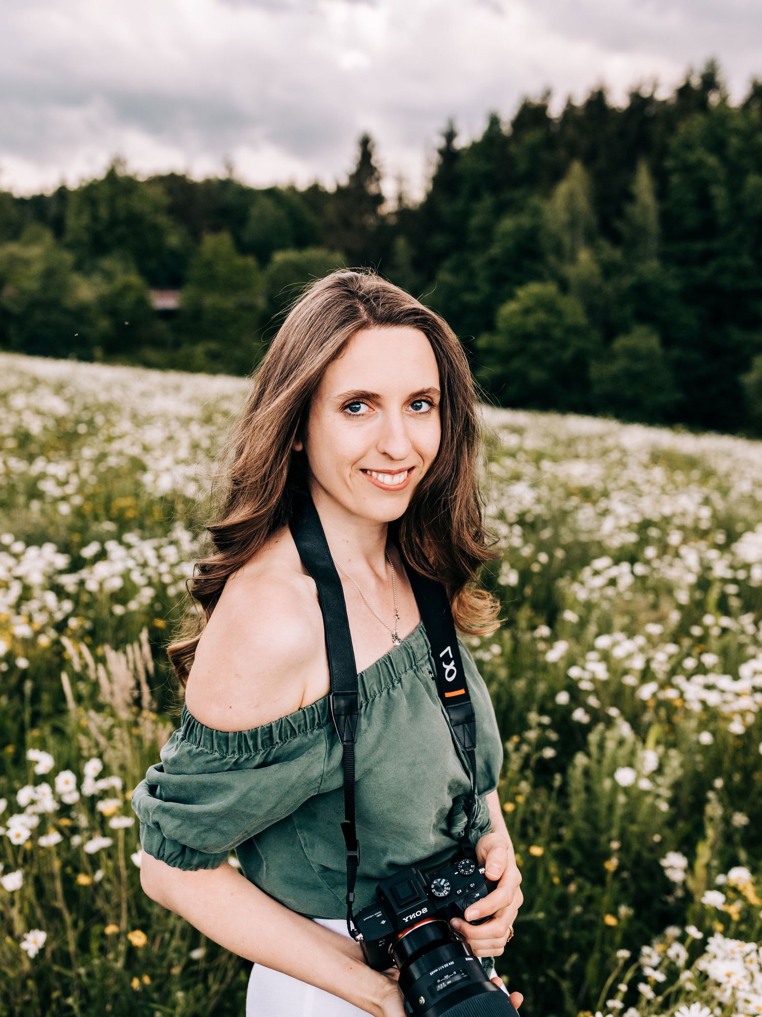 Portrait of photographer with camera in field of wildflowers under cloudy sky.