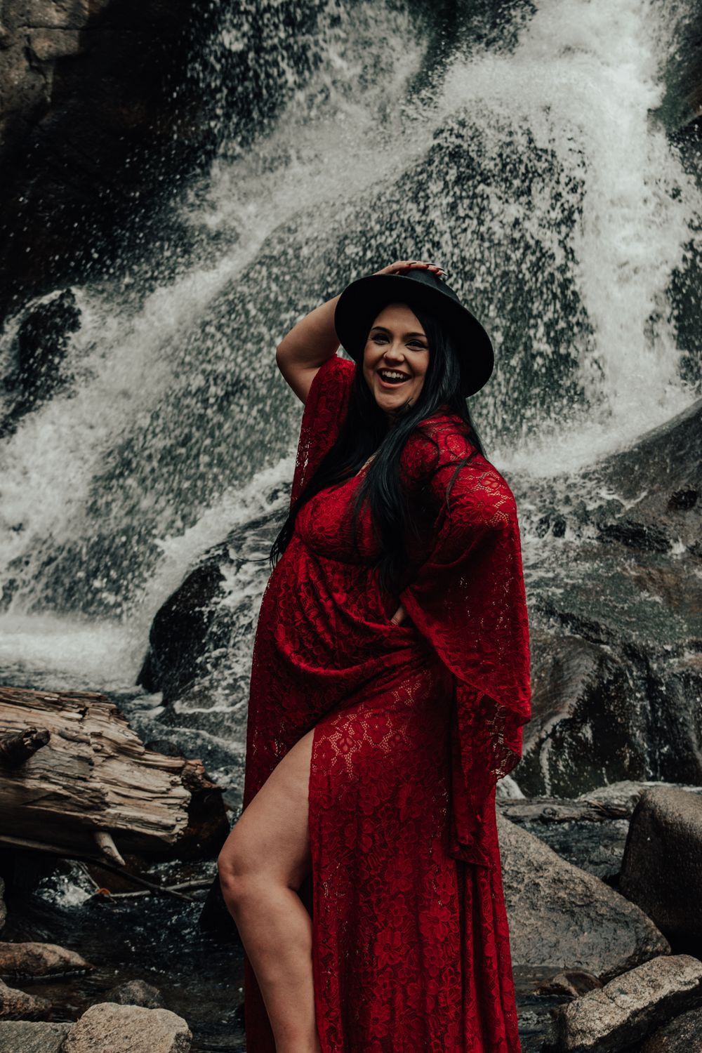 Smiling pregnant woman in red lace gown and black hat posing by waterfall.