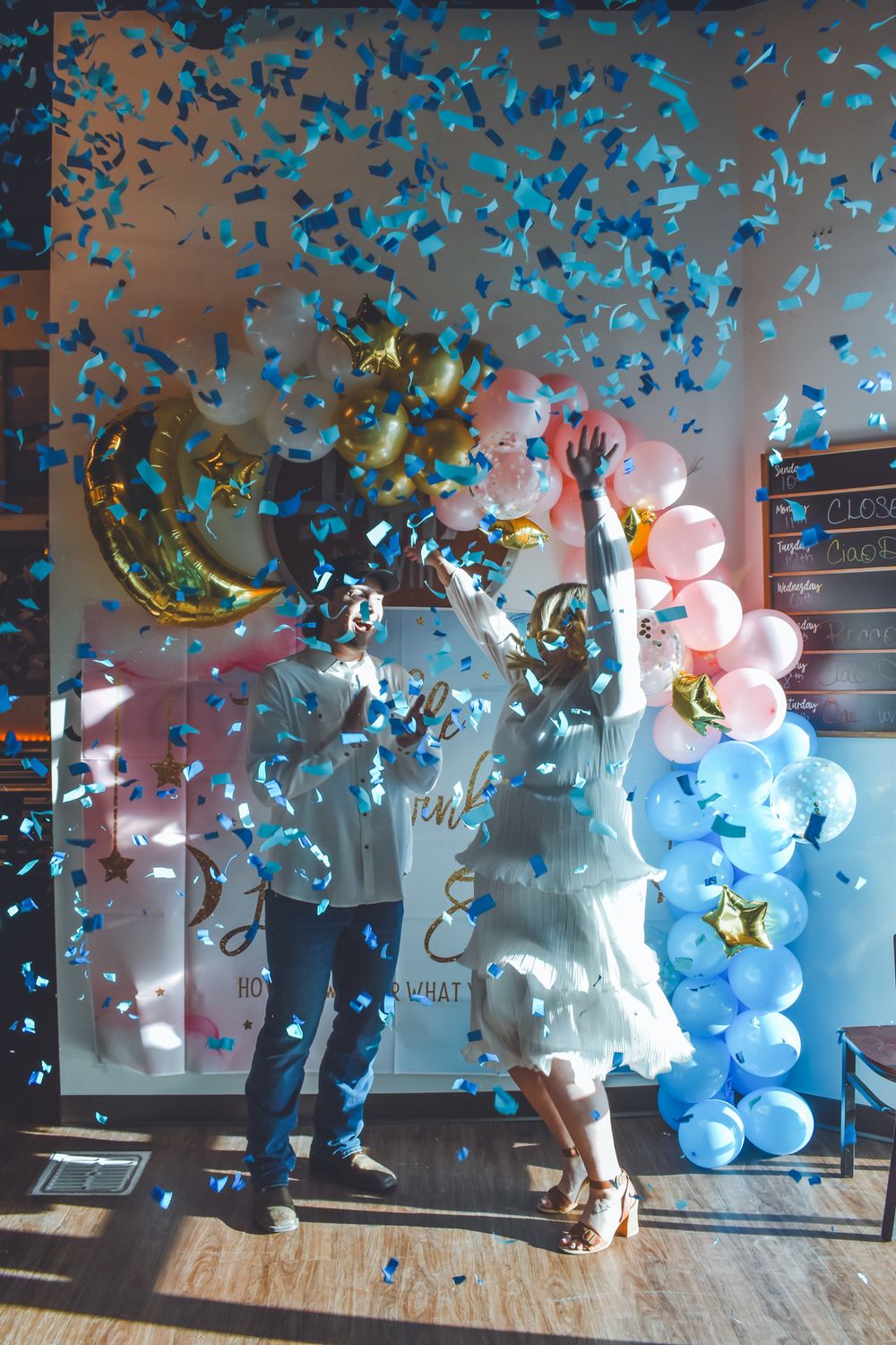 Couple celebrating gender reveal with blue confetti at a festive balloon-decorated party.