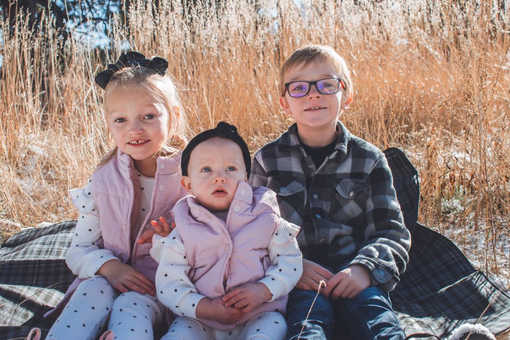 Three young siblings sitting together on a blanket in a golden autumn field.